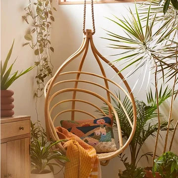 Hanging rattan chair with cushions and a blanket, surrounded by plants in a cozy room.