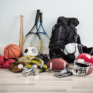 Collection of sports equipment including a basketball, soccer ball, football, baseball bat, tennis rackets, and backpack on a light wooden floor.